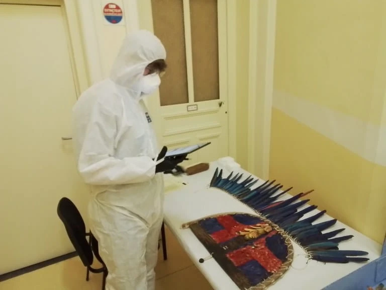 Técnico de macacão e máscara brancos examina um manto de plumas vermelhas e azuis sobre a mesa